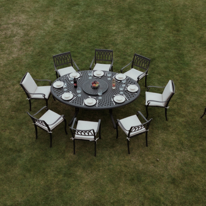 A circular outdoor dining table with eight chairs, each set with a white plate, surrounded by a green lawn and brick house.