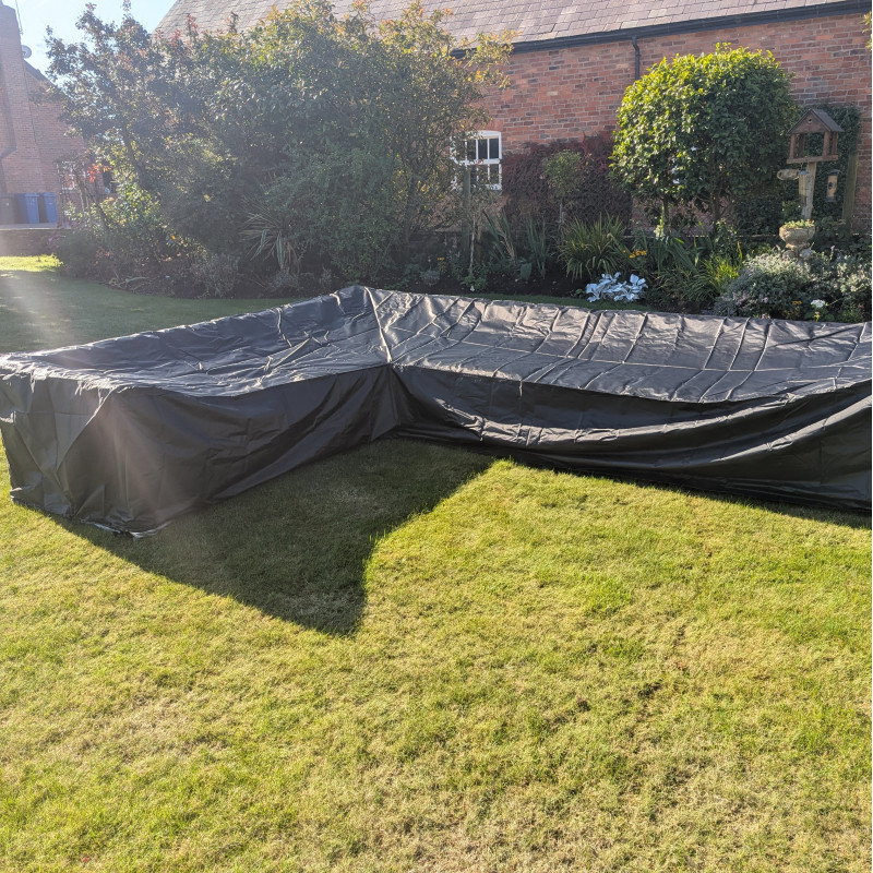 A large black tarpaulin covering a rectangular structure on grass, set against a garden background with trees and walls.