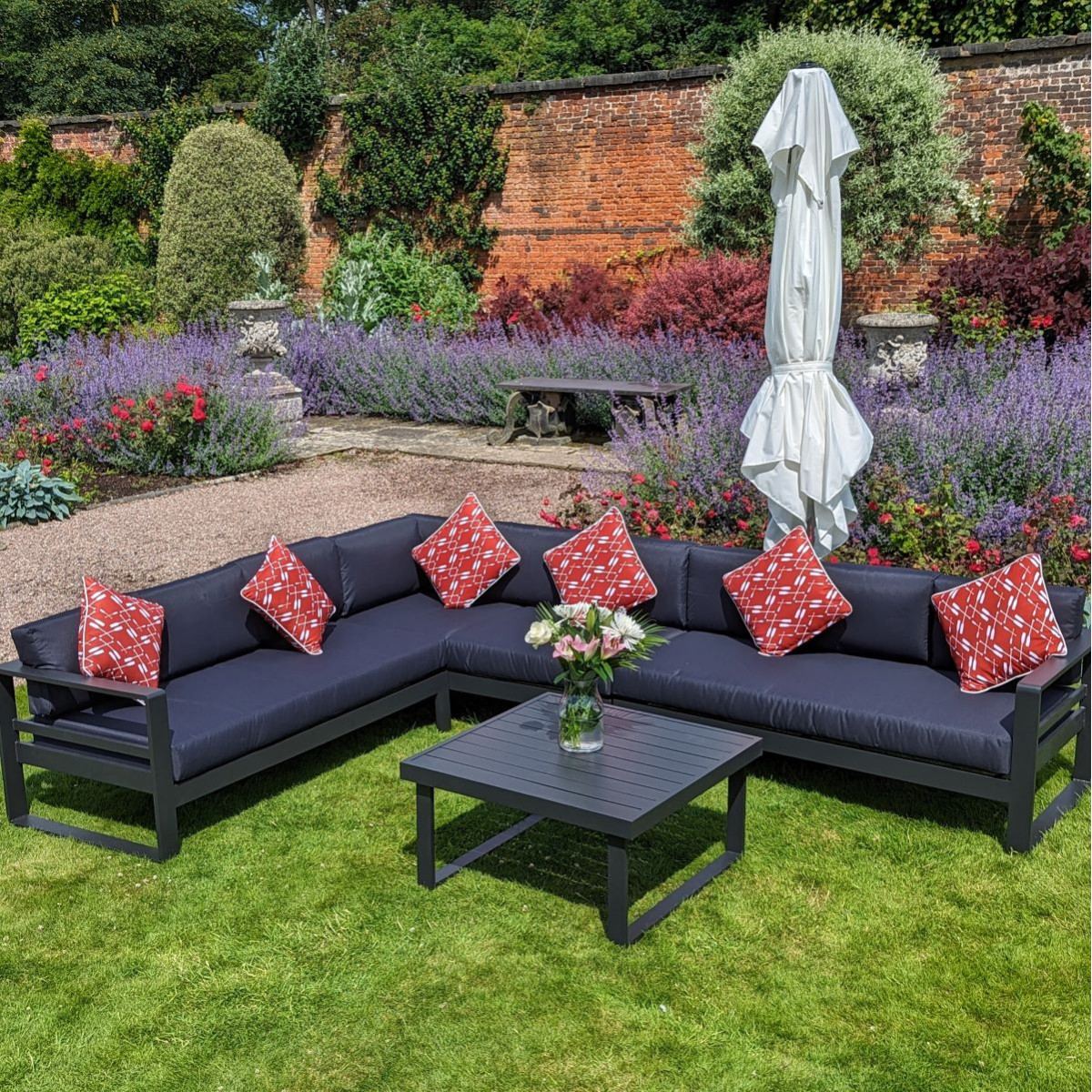 A modern outdoor seating area with a beige sofa, blue patterned cushions, and a table, shaded by a large white umbrella in a garden.