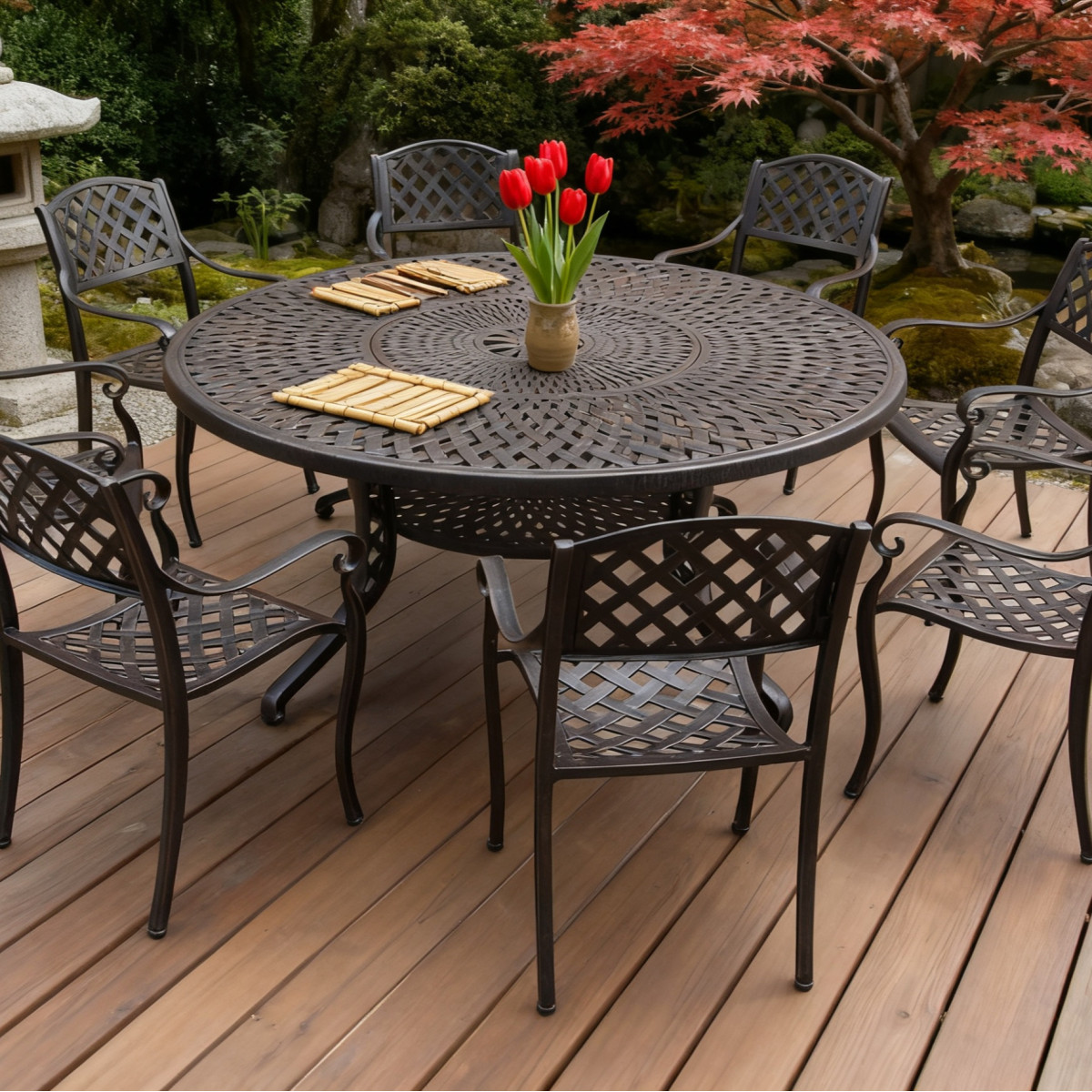 A round patio table with a vase of greenery, wine bottles, and fruit, set in a gravelled outdoor area near a brick house.