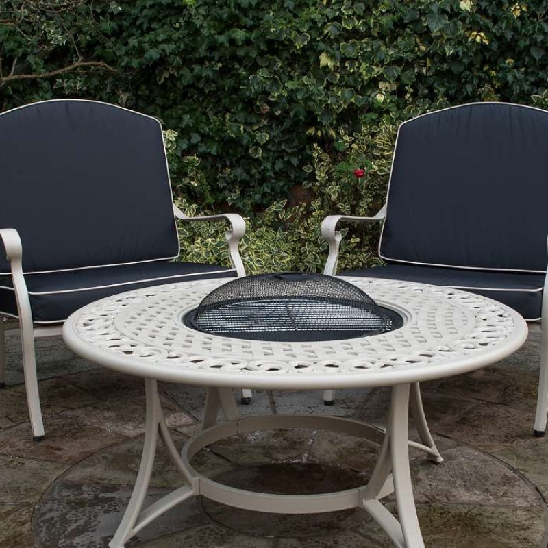 A black metal outdoor sofa with two cushions facing a round table featuring a circular design and a grill opening, set on gravel.