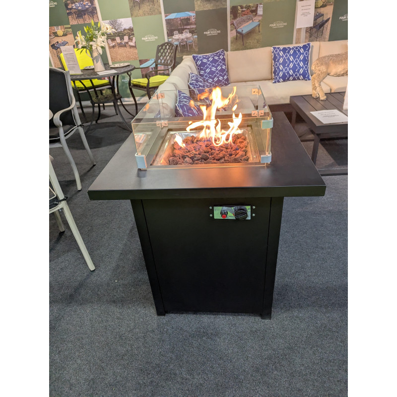 A black outdoor gas fire table with flames emerging from the top and control knobs visible on the front panel.