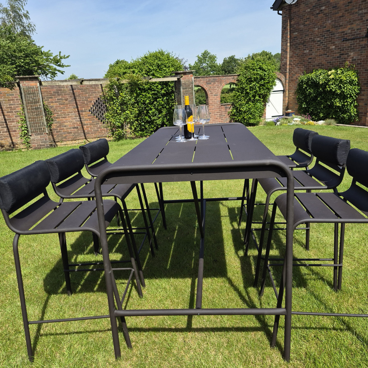 Outdoor table set with four bar stools, a bottle of red wine, and glasses, in a garden surrounded by greenery.
