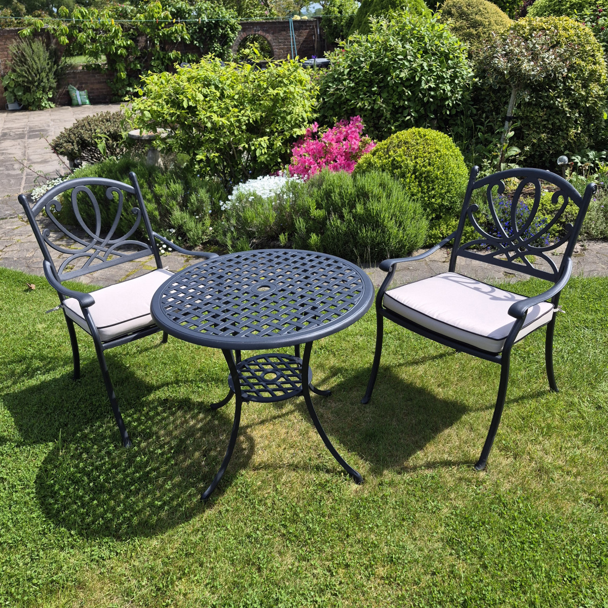 A small round black metal table surrounded by two chairs, set on green grass with a white brick wall and climbing ivy in the background.